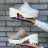 Two pairs of beige clogs held up against a brick wall.